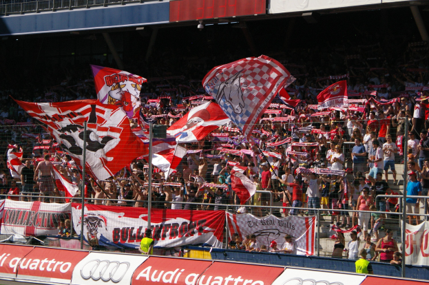 Eine große Menschenmenge in einem Stadion, einige halten Fahnen und Banner mit Text, andere stehen auf dem Boden, umgeben von einem Metallzaun, einem Schild, einem Anzeigebildschirm und einer Decke mit Deckenleuchten.