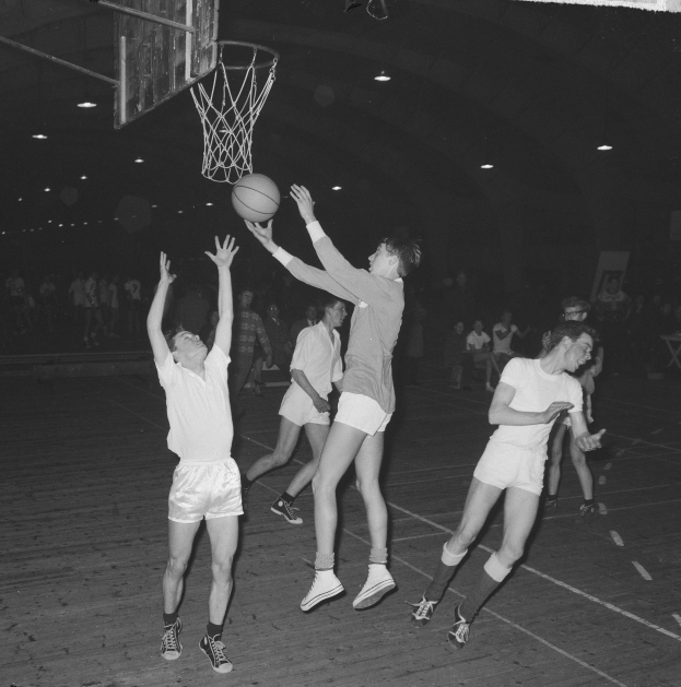 Schwarzes und weißes Foto von jungen Männern, die Basketball in einer Turnhalle spielen, mit einem Basketballkorb und Zuschauern im Hintergrund.