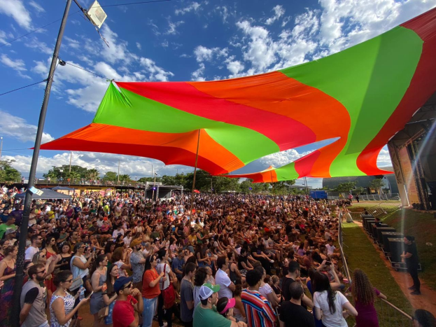 Eine große Menge steht vor einer Regenbogenflagge mit einem Gebäude, Lautsprechern auf der Bühne, Bäumen und Wolken im Hintergrund während des 10-jährigen Jubiläums eines Festivals.