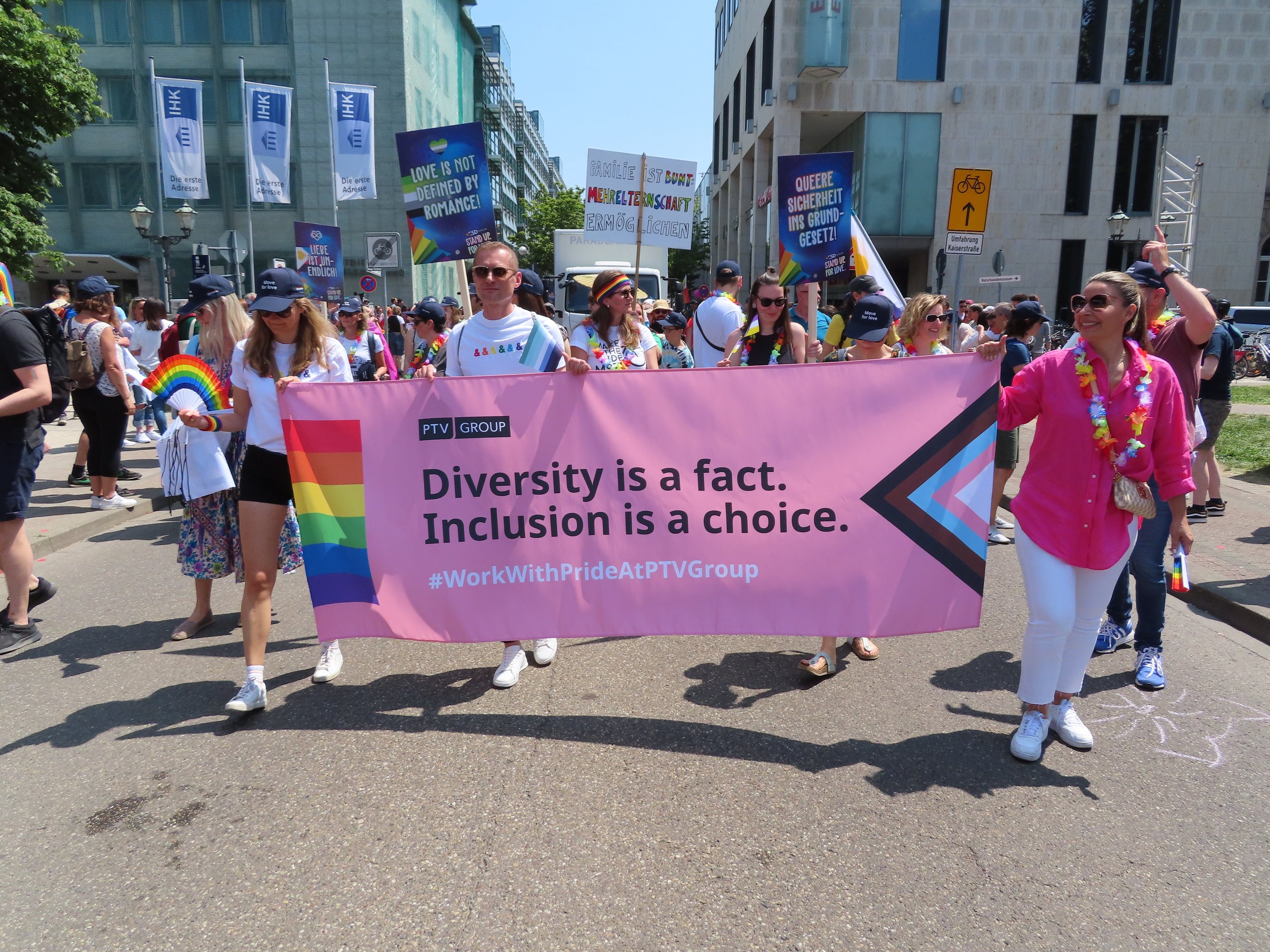 Eine Gruppe von Menschen marschiert auf einer Stra├če, h├Ąlt ein Banner mit der Aufschrift "Diversit├Ąt ist eine Tatsache, Inklusion ist eine Wahl" in den H├Ąnden, einige tragen K├Ąpfe und Schutzbrillen und andere halten zus├Ątzliche Banner, vor einem Hintergrund von Geb├Ąuden, B├Ąumen, Laternenmasten und einem klaren blauen Himmel.