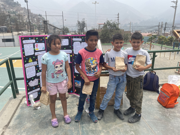 Gruppe von Kindern auf einem Basketballfeld mit Papiertüten in den Händen, Tüten und Gegenstände rechts daneben und eine Tafel mit Text links daneben, vor einem Hintergrund aus Maschendraht, Pfosten, Straßenlaternen, Bäumen, Gebäuden, Hügeln und Himmel.
