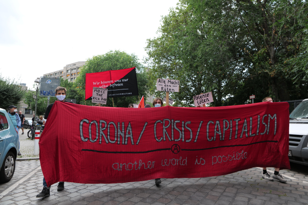 Eine Gruppe von Menschen marschiert mit einer roten Fahne, auf der 'Corona-Krise/Kapitalismus' steht, durch eine Straße, mit parkenden Fahrzeugen auf beiden Seiten und Gebäuden, Bäumen und einem klaren blauen Himmel im Hintergrund.