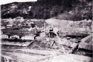 Schwarzes und weißes Foto eines Kohlebergwerks mit Gebäuden, Pfählen und Bäumen im Hintergrund, mit Text unten.