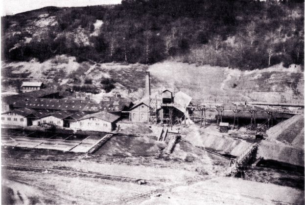 Schwarzes und weißes Foto eines Kohlebergwerks mit Gebäuden, Pfählen und Bäumen im Hintergrund, mit Text unten.