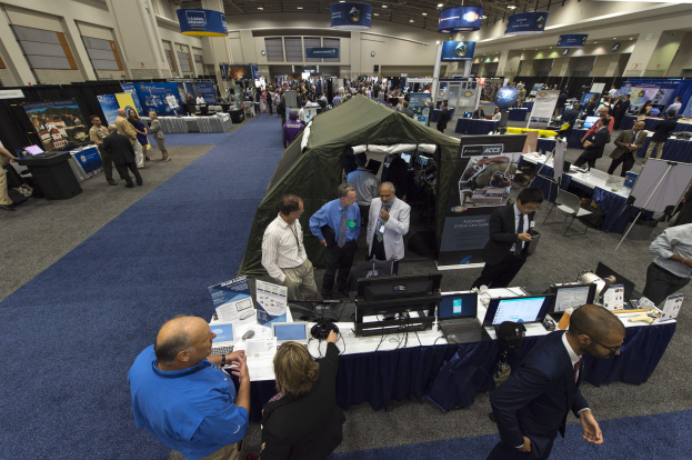 Eine große Gruppe von Menschen auf einer Karriere-Messe steht um Tische mit Laptops, Monitoren und Papieren herum, im Hintergrund ein Zelt, Schilder, Deckenleuchten und Fenster.