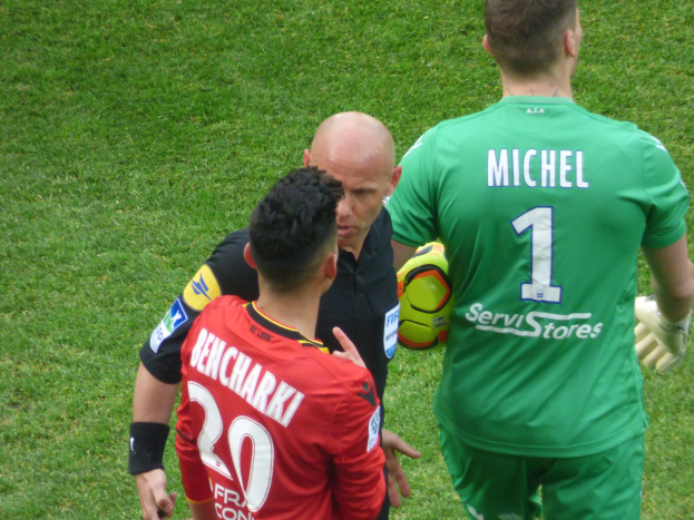Schiedsrichter spricht mit zwei Fußballspielern auf einem grünen Rasen, einer hält einen Ball.