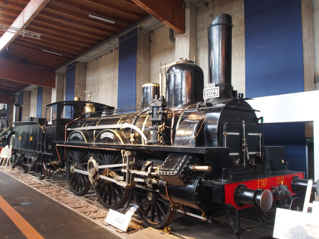 Alte Dampflokomotive in einem Museum ausgestellt, umgeben von Eisenbahnschienen, Informationsschildern und anderen Ausstellungsstücken unter einer beleuchteten Decke.