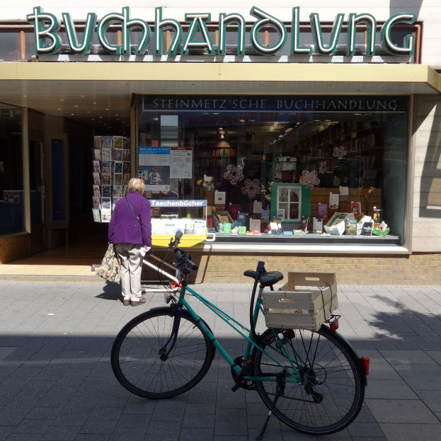 Eine Frau steht neben einem Fahrrad, das vor einem Geschäft geparkt ist, mit einem Fahrradrad im Vordergrund und Büchern, die durch die Glasscheibe des Geschäfts sichtbar sind.