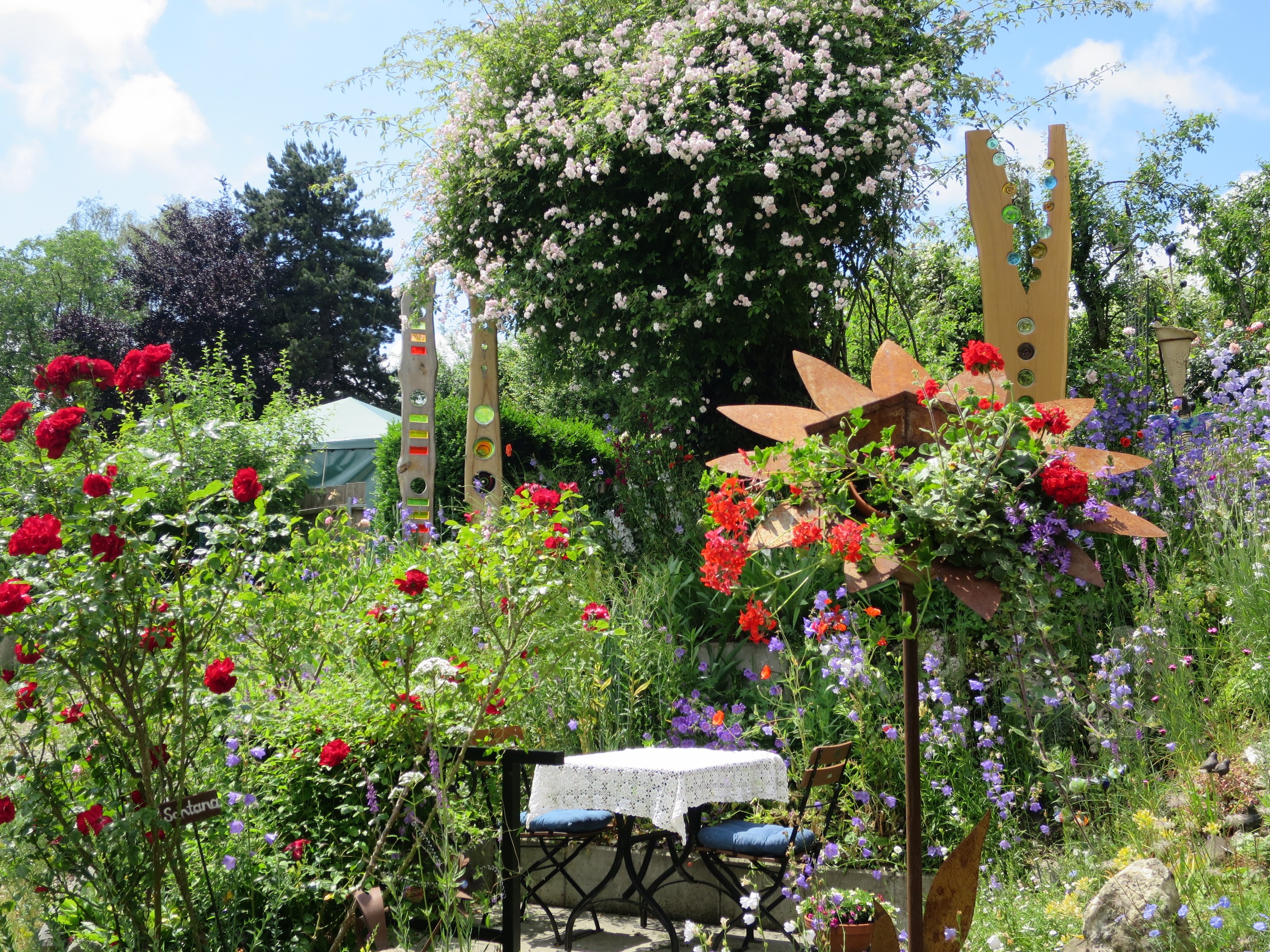 Ein Garten auf dem Chelsea Flower Show mit bunten Blumen und Pflanzen, einem Tisch mit einer Decke und Stühlen in der Mitte und Bäumen im Hintergrund unter einem bewölkten Himmel.