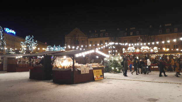 Ein verschneiter nächtlicher Weihnachtsmarkt mit Menschen, Buden, Pflanzen, Gebäuden und Schildern unter einem bewölkten Himmel.