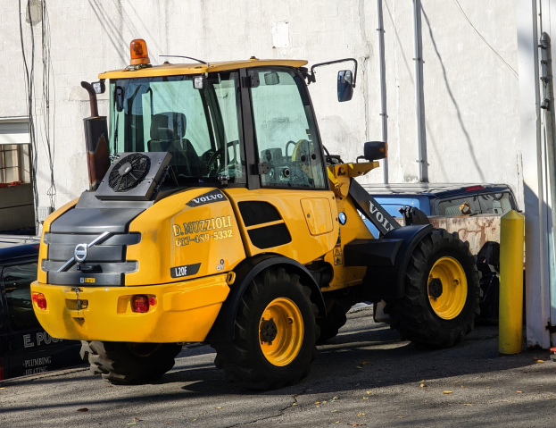 Ein gelber Radlader auf einer Straße vor einem Gebäude mit einer Wand im Hintergrund und anderen Fahrzeugen in der Nähe.