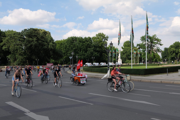 Eine Gruppe von Menschen, die auf Fahrrädern eine von Bäumen gesäumte Straße mit Laternenmasten und Fahnenmasten entlangfahren, während des Pariser Fahrradrennens 2016, mit bewölktem Himmel und einigen Fahrzeugen im Hintergrund.