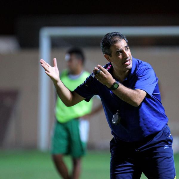 Ein Mann in einem blauen T-Shirt und Hose steht auf einem Fußballfeld und hält ein Mikrofon in der Hand, während er gestikuliert. Im Hintergrund sind eine Person in einem grünen Kleid, ein Torpfosten und eine Wand zu sehen.