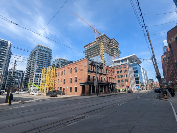 Eine belebte Straße in Toronto mit fahrenden Fahrzeugen, Füpßlern auf dem Gehweg, Strommasten mit Drühten, Schildern, Geböuden und einer entfernten Baustelle mit einem Kran gegen einen bewölktem Himmel.