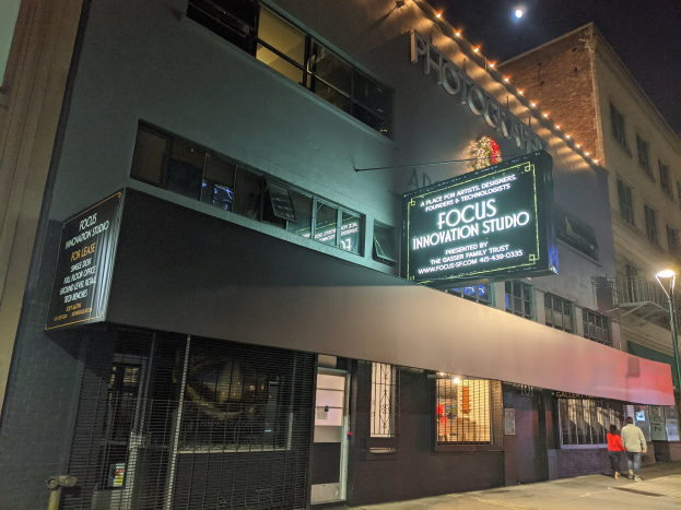 Außenansicht des Focus Innovation Studio in New York City bei Nacht, mit beleuchteten Fenstern und Namensschildern an umliegenden Gebäuden, Menschen auf dem Bürgersteig und dem Nachthimmel im Hintergrund.