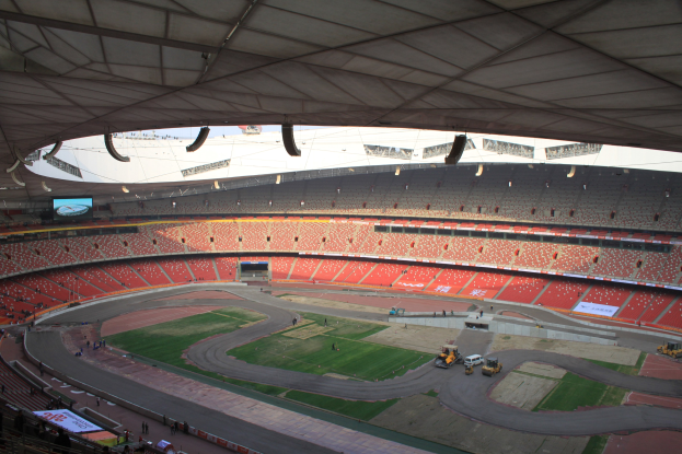 Großes Stadion voller Zuschauer, umgeben von Fahrzeugen, Rasen, Texttafeln, Geländern, einem Bildschirm und Deckenbeleuchtung.