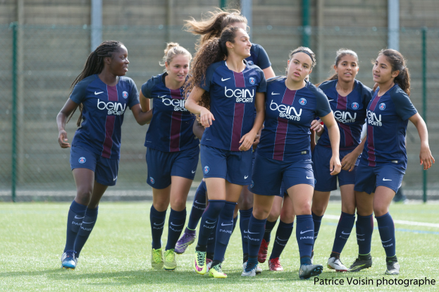 Gruppe junger Frauen beim Fußballspielen auf einem Rasenfeld mit Maschendrahtzaun und einer Wand im Hintergrund, Text in der rechten unteren Ecke lautet "Paris Saint-Germain Frauenfußball"