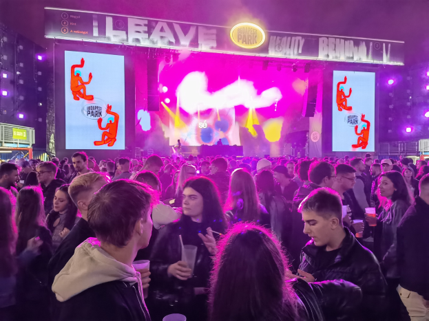 Eine große Menge steht vor einer hell erleuchteten Bühne, umgeben von Tafeln mit Text und Bildern, wobei einige Teilnehmer Gläser halten.