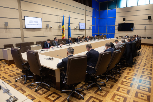 Eine große Gruppe von Menschen sitzt um einen langen Tisch mit Mikrofonen und Papieren in einem Raum mit Flaggen der Europäischen Union, Fernsehern, Deckenleuchten und Lautsprechern, was auf eine Sitzung des Europäischen Parlaments in Brüssel hinweist.