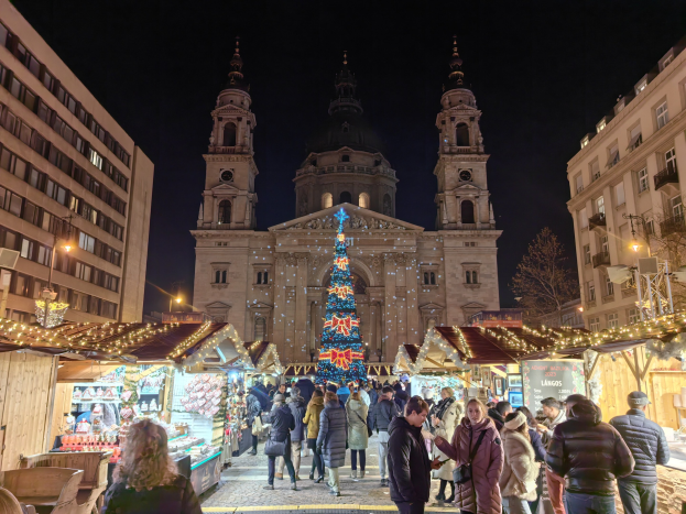 Ein belebter Weihnachtsmarkt vor einer Kirche bei Nacht, mit Menschen um beleuchtete Stände, Gebäude, Bäume und einen sternenklaren Himmel.