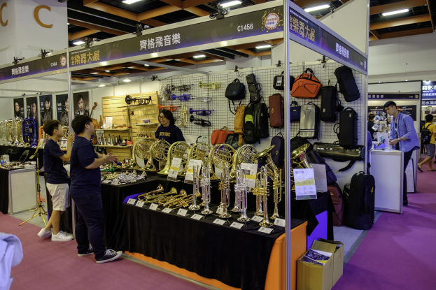 Eine Gruppe von Menschen um einen Tisch mit Musikinstrumenten, Taschen und anderen Gegenständen beim Hongkonger Internationalen Musikfestival, mit Schildern, Deckenbeleuchtung und einem Karton auf dem Boden.