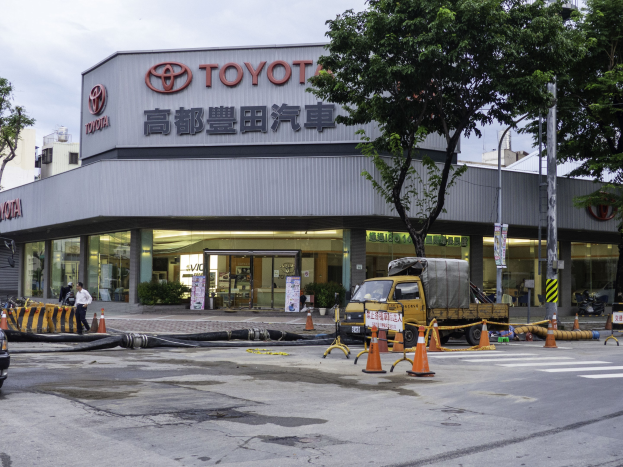 Ein Toyota-Autohaus in Tokio, Japan, mit Fahrzeugen, Verkehrskegeln, Rohren, Menschen, Bäumen, Pfählen, Planken, Gebäuden und einer bewölkten Himmel.