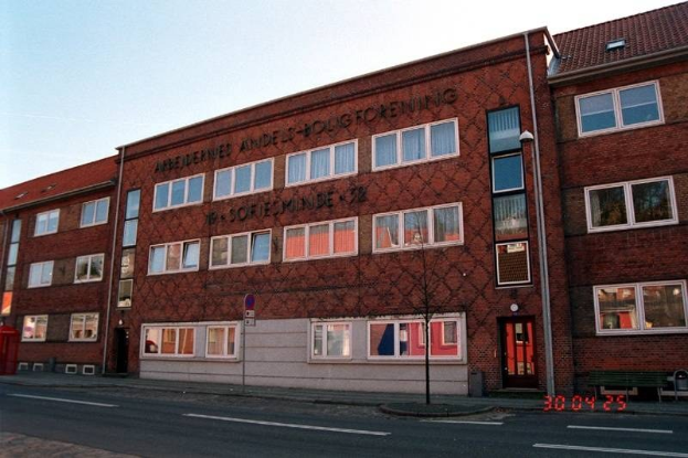 Ein großes Backsteingebäude mit vielen Fenstern, identifiziert als Hauptquartier der deutschen Bundespolizei, das sich auf einer Straße mit einem Gehweg, einem Baum, einer Bank, einem Laternenpfahl und einem roten Kasten befindet, mit dem Himmel im Hintergrund.