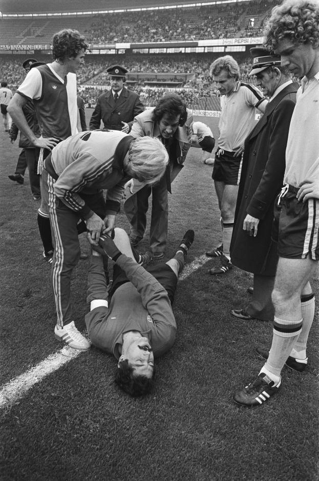 Schwarzes und weißes Bild eines Fußballspielers, der auf dem Boden liegt und von einer Gruppe von Männern umgeben ist, mit einer Menge im Hintergrund des Stadions.