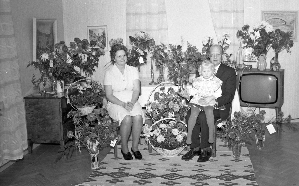 Schwarzes und weißes Foto einer Familie aus drei Personen - ein Mann hält ein Kind, eine Frau und das Kind - sitzen auf einem Sofa im Wohnzimmer mit Blumenvasen, einem Teppich, gerahmten Wanddekorationen und Vorhängen.