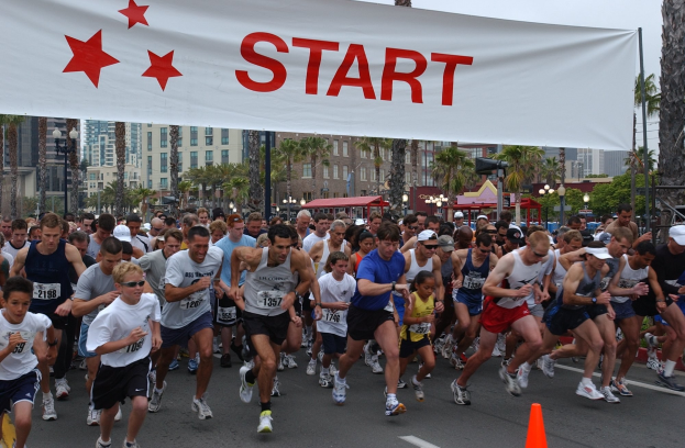 Gruppe von Läufern bei einem Marathon, die an einer Verkehrsinsel vorbeilaufen, mit einem Banner und Gebäuden im Hintergrund unter einem klaren Himmel.