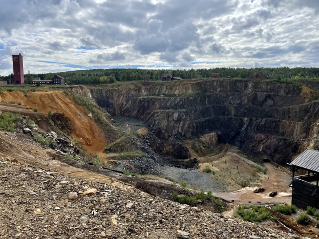 Großer Tagebau in einem Schmutzfeld umgeben von Bäumen, Gebäuden und einem Schuppen unter einem bewölkten Himmel, mit verstreuten Steinen und Pflanzen.
