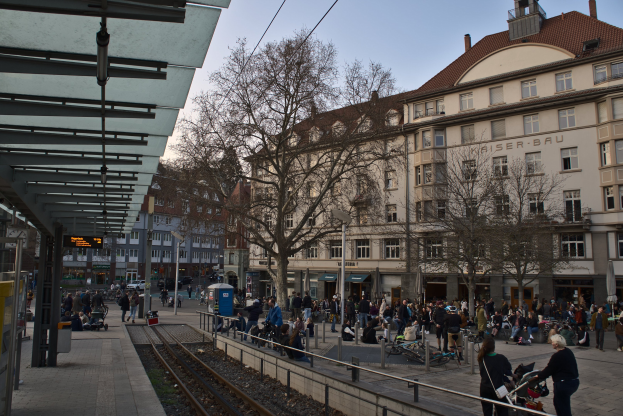 Eine belebte Stadtstraße mit einem Bahnhof auf der linken Seite, umgeben von Gebäuden, Bäumen, Laternen und Texttafeln, wo Menschen gehen, auf Bänken sitzen oder stehen, unter einem sichtbaren Himmel.