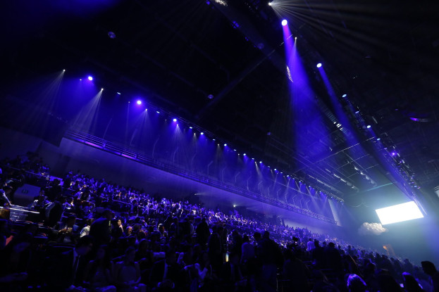 Große Menschenmenge in einem schwach beleuchteten Konzertsaal, der einer Bühne mit Deckenleuchten und einem Bildschirm auf der rechten Seite gegenüberliegt.