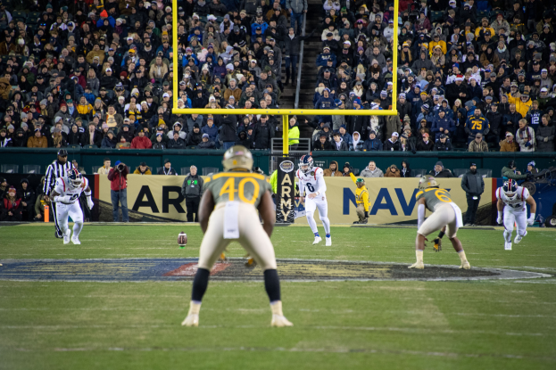 Fußballspieler in Sportkleidung und Helmen konkurrieren auf einem Feld mit Zuschauern in den Rängen, Polen und Tafeln mit Text sichtbar.