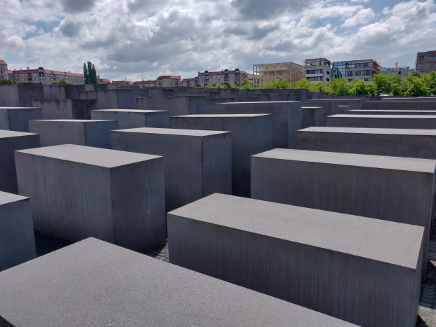Konkreter Holocaust-Denkmal mit rechteckigen Blöcken, die in einem Gitter angeordnet sind, umgeben von Bäumen, Gebäuden und einem bewölkten Himmel.