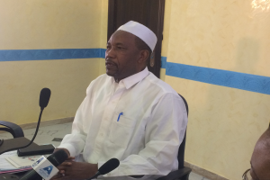 Ein Mann in einem weißen Kleid und einer Mütze sitzt an einem Tisch mit Mikrofonen und spricht, während Papiere und Gegenstände auf dem Tisch zu sehen sind, mit einer anderen Person mit Brille rechts von ihm, in einem Raum mit einer Wand und einer Tür im Hintergrund.