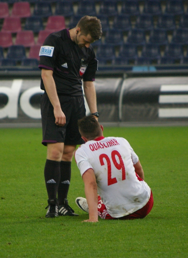 Ein Fussballspieler in Sportkleidung sitzt neben einem Schiedsrichter auf dem Boden in einem Stadion mit Plakaten und Stühlen im Hintergrund.