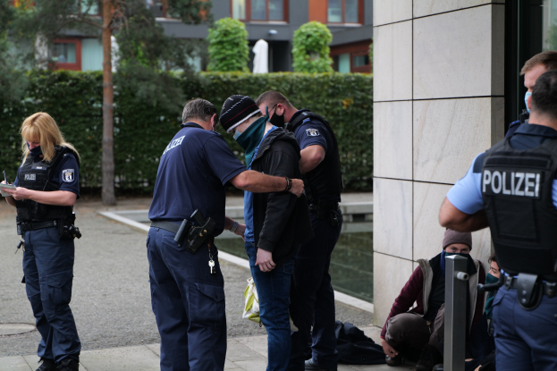 Deutsche Polizeibeamte vor einem Gebäude mit einer Frau, die ein Buch und einen Stift hält, während andere in der Nähe sitzen oder stehen, mit Bäumen und Gebäuden im Hintergrund.