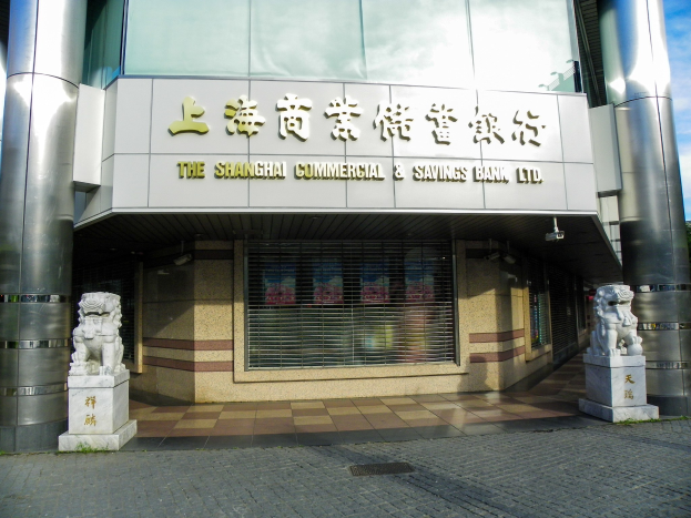 Das Bild zeigt das Shanghai Commercial & Savings Bank Ltd.-Gebäude mit Säulen und einem Namensschild, flankiert von Statuen, vor einem Baum- und wolkenverhangenen Himmel.