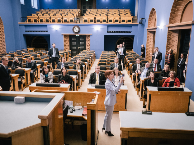 Frau hält Rede im schottischen Parlament, vor sitzenden und stehenden Zuhörern mit Mikrofonen, Gläsern, Papieren und anderen Gegenständen auf Tischen, mit einer Uhr und Deckenbeleuchtung im Hintergrund.