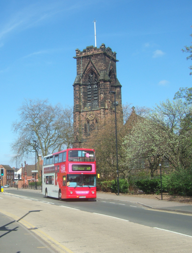 Ein roter Doppeldeckerbus fährt auf einer Straße neben einer Kirche, mit Gebäuden, Bäumen, Pflanzen, Laternen, Verkehrszeichen, anderen Fahrzeugen und einer bewölkten Landschaft im Hintergrund.