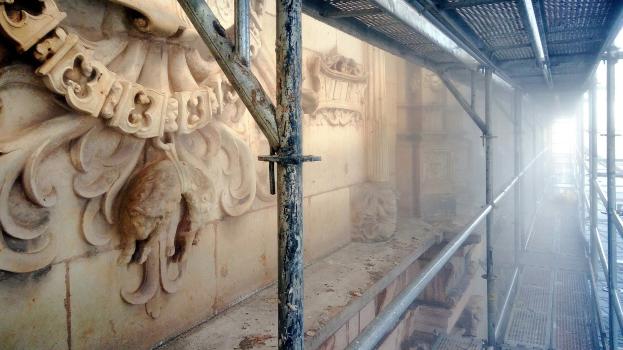 Ein renoviertes Gebäude mit detaillierten Schnitzereien an der linken Wand, Metallstangen an der rechten und Gerüst drumherum.