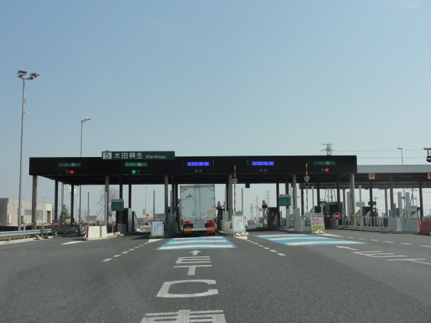 Mautstelle mit vor der Mautstelle geparktem Lastwagen, stra├čenrand mit wei├čen und blauen Markierungen, Laternenmasten und Schildern auf beiden Seiten, Geb├Ąude und B├Ąume im Hintergrund unter einem klaren blauen Himmel.