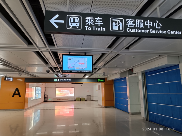 Innenansicht eines Bahnhofs mit einem "Zum Bahnkunden-Service-Zentrum"-Schild oben, sichtbarer Boden, Türen auf der rechten Seite, Deckenanzeigen, ein zentraler Bildschirm und ein Wasserzeichen unten rechts.