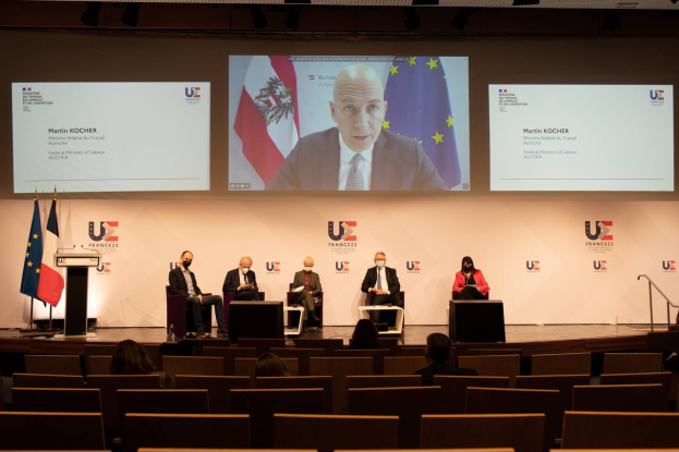 Eine Gruppe von Menschen sitzt auf einer Bühne in einem Halbkreis um ein zentrales Podium mit Mikrofon, gewandt einem Publikum zu, mit Flaggen auf beiden Seiten und einem Hintergrundbild, beleuchtet von Deckenlampen während einer Pressekonferenz der Europäischen Union.