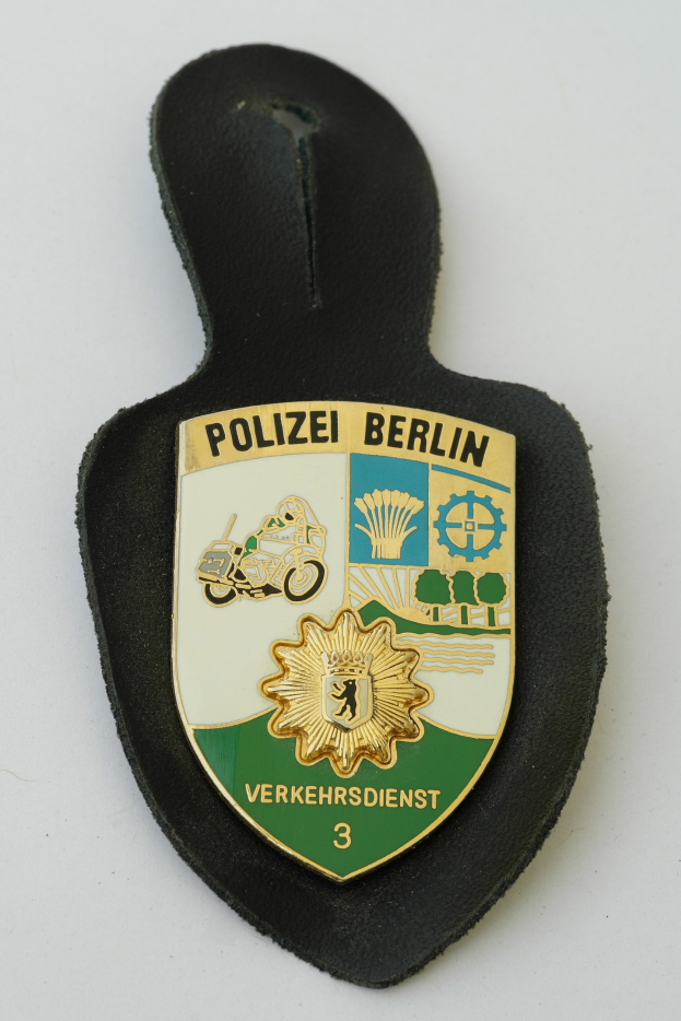 Ein schildförmiges Polizeibadge mit "Polizei Berlin" in schwarzer fettgedruckter Schrift auf einem blauen Hintergrund mit einem weißen Stern und Rahmen, das auf einer Oberfläche ruht.