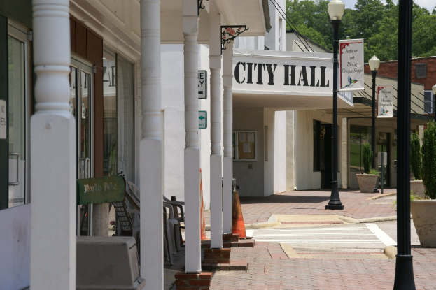 Rathaus mit umliegenden Gebäuden, Straßeninfrastruktur einschließlich Masten und Laternen, Fahrzeugen, Verkehrskegeln, Außenmöbeln, Pflanzen, Bäumen und einem bewölkten Himmel.