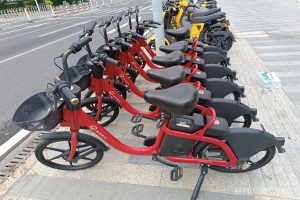 Eine Reihe roter und schwarzer E-Bikes, die an einer Straße mit einem weißen Pfahl im Vordergrund, Bäumen, Gebäuden und einem klaren blauen Himmel im Hintergrund geparkt sind.