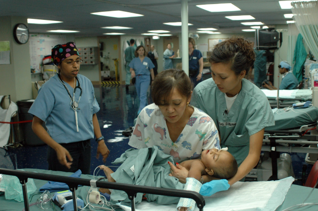 Eine Gruppe von medizinischen Fachleuten in Kitteln versammelt sich um ein Krankenhausbett mit einem Baby, während eine Frau das Kind hält und andere Stethoskope tragen.