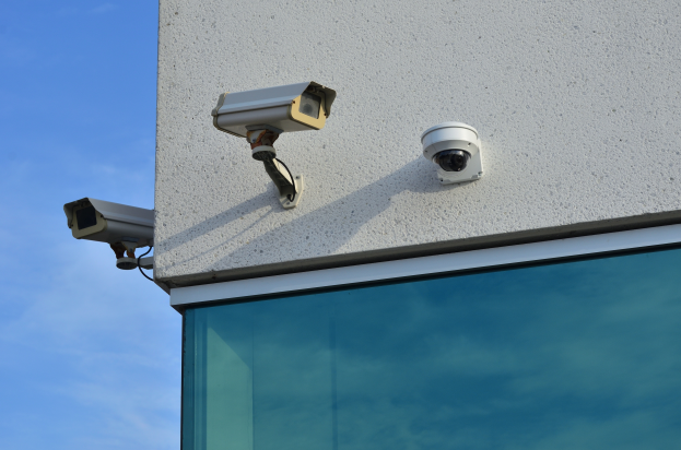 Zwei Überwachungskameras an der Seite eines Gebäudes montiert, mit einer Glaswand im Vordergrund und einem klaren blauen Himmel im Hintergrund.